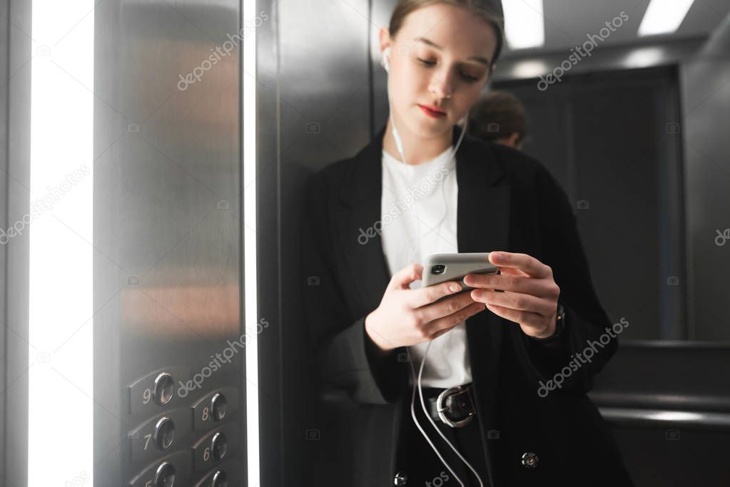 Close up photo of businesswoman holding smartphone in her heands in the elevator and listening to music in headphones. Portrait of female office worker's hands with smartphone and headphones in lift.