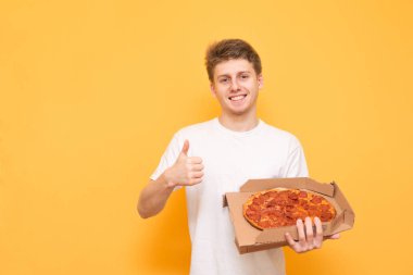 Beyaz bir gömlek gülümseyen adam elinde bir pizza kutusu ile ayakta, parmağını gösterilmesini ve sarı bir arka plan üzerinde kameraya bakıyor. Pizza kavramı.