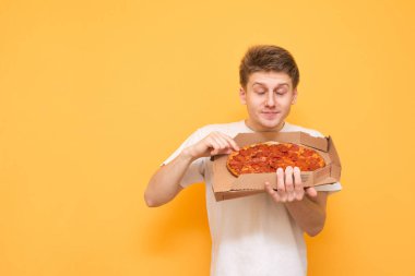 Komik genç adam bir kutu taze pizza tutar ve aroma sahiptir, sarı bir arka plan üzerinde pizza burnunu çekmek. Adam bir kutu sarı bir arka plan üzerinde sıcak pizza kokluyor. Boşaltmak