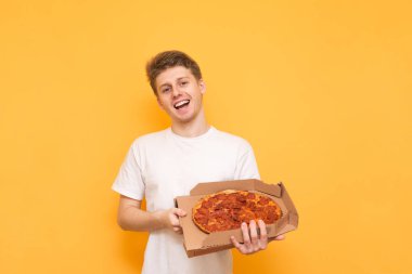 Pizza içine belgili tanımlık fotoğraf makinesi ve gülümsüyor arıyorum onun elinde bir kutu ile sarı bir arka plan üzerinde mutlu genç adam. Pizza teslim üzerinde tutarak ve gülümseyen, beyaz bir T-shirt giyen genç adam izole