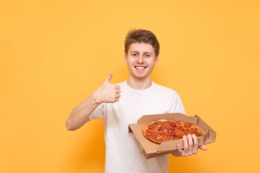 Kamera ve gösteren parmak mutlu genç adam pizza onun elinde bir kutu ile sarı bir arka plan. Bir beyaz adam gülümseyerek T-shirt bir pizzacı elinde tutan ve bir jest gösterir