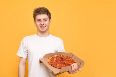 Pozitif adam bir beyaz T-shirt, sarı bir arka plan üzerinde izole bir kutu pizza onun elinde tutan, içine belgili tanımlık fotoğraf makinesi ve gülümsüyor görünüyor. Fast Food kavramı.