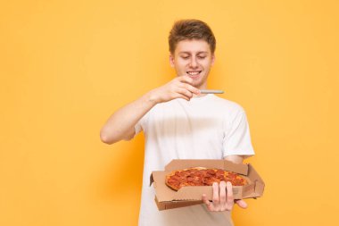 Gülümseyen adam bir kutu taze pizza tutar ve bir smartphone cep telefonu ile sarı bir arka plan üzerinde izole, bir fotoğraf yapar. Mutlu genç adam bir pizza sarı bir arka plan üzerinde fotoğraf çekimi.