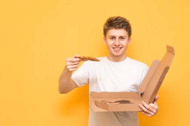 Mutlu gülümseyen genç bir kutu tutar ve bir parça pizza onun elinde, kameraya görünüyor ve sevinir, izole. Beyaz bir t-shirt giyen sarı bir arka plan üzerinde elinde bir pizza ile.