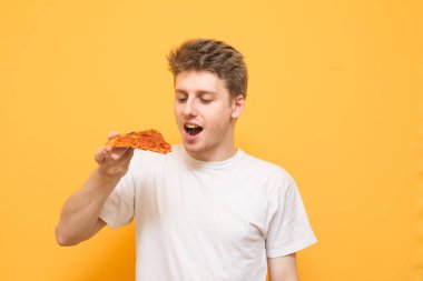 Bir parça pizza sarı bir arka plan üzerinde izole onun elinde olan aç bir adam portresi. Genç adam sarı bir arka plan üzerinde duruyor ve elinde bir parça pizza üzerinde odaklanmıştır.