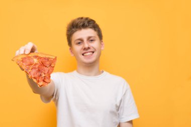 Neşeli genç adam sarı bir arka plan üzerinde bir kamera bir parça iştah açıcı pizza gösterir. Bir parça pizza onun elinde, yakın çekim ve odak ile gülümseyen adam. Arka plan. Boşaltmak