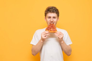 Beyaz tişört ve bir parça pizza genç adam sarı bir arka plan üzerinde izole. Yakışıklı çocuk sarı bir arka plan üzerinde duran bir parça iştah açıcı pizza yiyor.