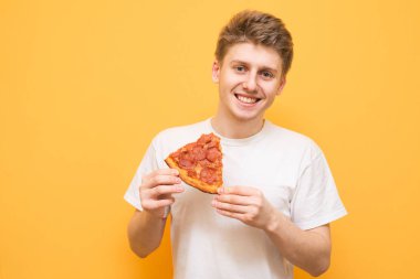 Gülümseyen bir adam portresi bir parça pizza onun elinde tutan ve kameraya bakarak izole sarı bir arka plan üzerinde beyaz tişört giymiş. Genç ve fast food. Boşaltmak