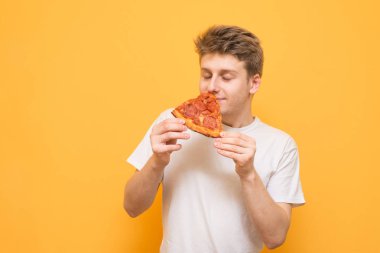 Genç adam tutar bir parça bir koku gözleriyle kokmaya elinde taze pizza, sarı bir arka plan üzerinde izole kapalı. Adam lokanta sarı bir arka plan üzerinde sahiptir.