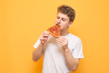 Beyaz bir T-shirt giyen sarı bir arka plan üzerinde kapalı gözleriyle iştah açıcı bir parça pizza yiyor ve zevk alır. Genç adam sarı bir arka plan üzerinde izole pizza, ısırıkları.