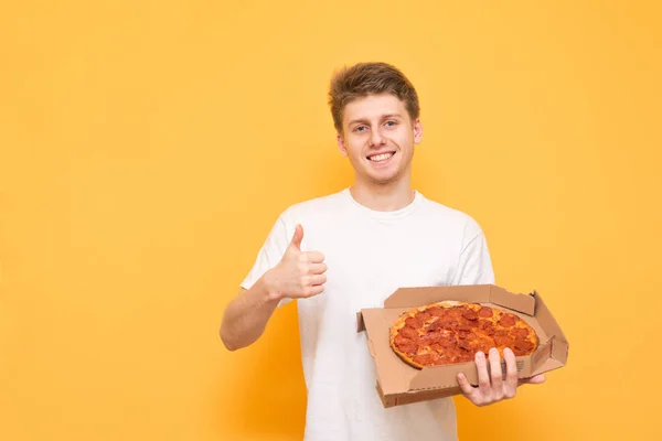 Smiling guy in a white t-shirt is standing with a box of pizza in his hands, showing his thumb up and looking into the camera on a yellow background. Pizza Concept.