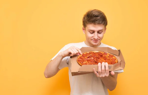 シャツを着た男は 黄色の背景の上に立って 空腹そうな顔と彼の手でピザの箱を見えます 黄色の背景で隔離ボックスで空腹の若い男 — ストック写真