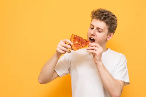 一个年轻人在黄色背景上吃了一块新鲜美味的比萨饼的肖像 饥饿的家伙在他的手中一块比萨饼 看着快餐 — 图库照片