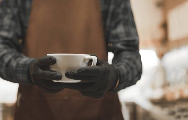 Bir fincan kahve barista yakın çekim elinde. Bir önlük barista ve siyah eldivenleri bir fincan kahve elinde, bir yakın çekim fotoğraf tutar. Boşaltmak