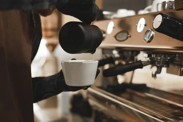 Barista pouring milk in to coffee, hot drink, close up view. Background photo of coffee shop owner making coffee by pouring milk in a cup.