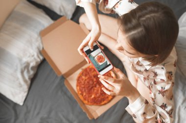 Bir smartphone cep telefonu ile pijama bir yatakta otururken, bir pizza kutusu resmini yapan bir kız portresi. Kadın yatakta bir pizza kutusu fotoğraf çekme. Kız ve yatakta kahvaltı için pizza.