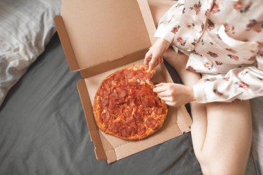 Yakın çekim fotoğraf bir pijama içinde bir kız yatakta oturur ve bir parça pizza kutusundan alır. Kızın bacakları ve pizza, bir kutu alır eller iştah açıcı bir parçası. Yatakta pizza yemek.