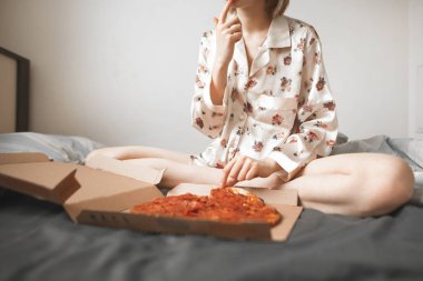 Girl in a pajama sits on the bed in the bedroom and eats a pizza from the box. Cropped photo of a woman with a box of pizza on the bed. Fast Food, Pizza Delivery. clipart