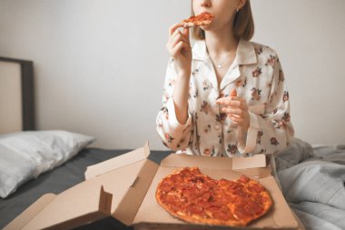 Güzel kız pijama giymiş rahat bir yatak odasında yatağın üstüne oturan ve pizza kutusundan yemek, bir parça ısırma ve zevk. Bir gün izin günü kahvaltıda yatakta sıcak pizza. Pizza teslim