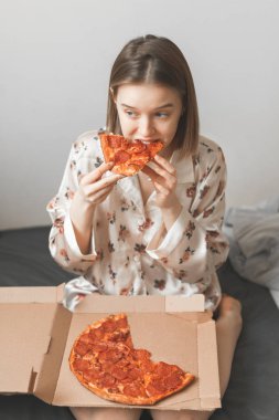 Bir pijama kız yatak odasında yatağın bir kutu onun kucağına oturur ve iştah açıcı bir parça pizza yiyor. Kadın yatakta pizza yiyor. Dikey fotoğraf. Boşaltmak