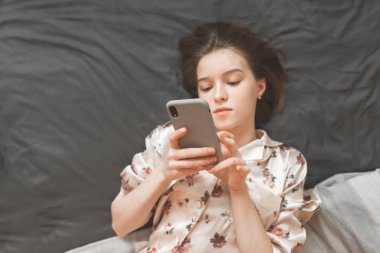 Pijama konsantre kız evde yatağım, bir akıllı telefon elinde tutan ve internet, yukarıdan bir görünüm kullanır. Smartphone üzerinde oturan bir kız eline odaklanın