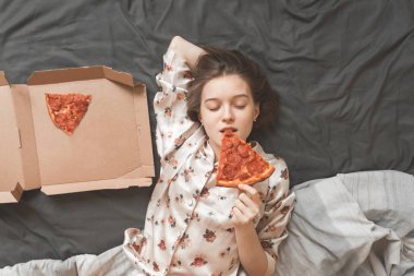 Güzel bir kız portresi pijama giymiş, gözleri kapalı bir parça pizza ısırıkları ve zevk alır. Tatlı kız evde istirahat, yatakta bir pizza yemek ve zevk. Üstten Görünüm.
