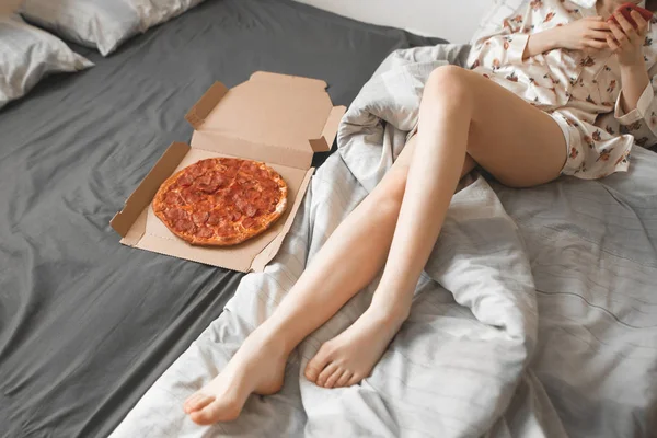 Slender girl in a pajama lays in a bed with a pizza box and uses a smartphone. Girl and pizza box for breakfast. Beautiful legs and a box with a fresh pizza on the bed.