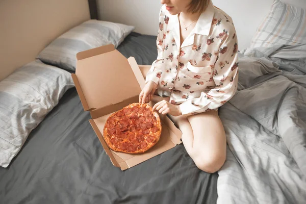 Pretty woman in a pajama sits on a bed with a pizza box and takes a piece. Girl sleeps pizza on the bed. Fast Food Concept.