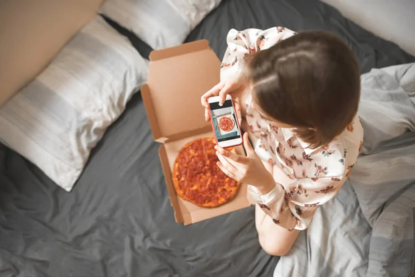 Lady in a pajama sits on a bed with a box of pizza and makes a photo on a smartphone. Girl picks up a breakfast, a box of pizza from the delivery. Photo of a pizza on a smartphone camera.