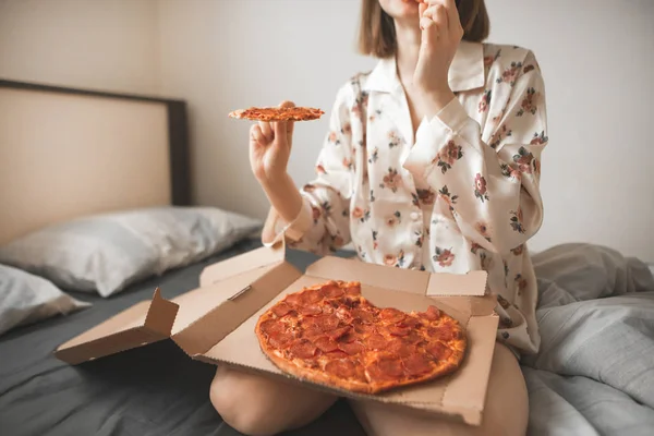 女の子のパジャマを着て ベッドに座ってピザのボックスで一枚と彼女の膝の上彼女の手で 少女をベッドの上配信で朝食ピザ食べる 写真をクローズ アップ — ストック写真