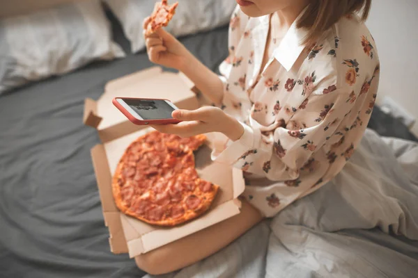 クローズ アップは パジャマを着て ベッドでピザを食べて スマート フォンを使っての少女の写真をトリミングしました 女性がピザの箱が付いているベッドに座って食べるファーストフード スマート フォンにメッセージを書く — ストック写真