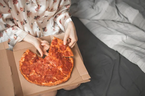 パジャマの女の子は寝室でベッドの上に座っている クローズ アップ ボックスや手 ピザからあつあつのピザの部分がかかります 女性は ベッドの上にピザを食べる Copyspace — ストック写真