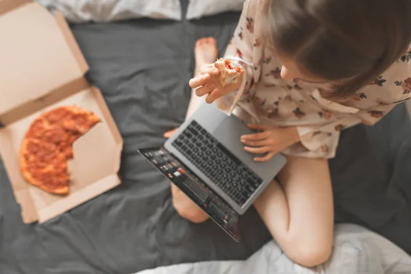 Portrait of a girl sitting in a bed with a box of pizza and using the Internet on a laptop, holds a piece of pizza in her hand. Work and breakfast in bed. Fast Food Concept. Pizza delivery.
