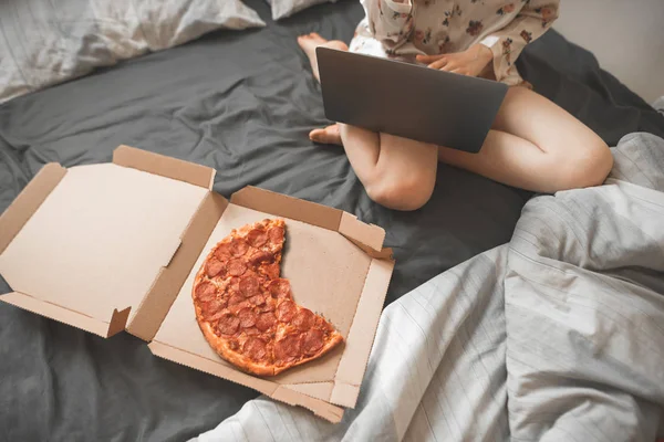 女の子がピザの箱の部屋にホーム パジャマ座っているであり ラップトップを使用しています 若い魅力的な女性フリーランサーが自宅のベッド上で動作し ピザを食べる — ストック写真