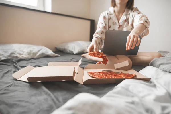 パジャマの女の子ホーム ベッドの上に座って ラップトップ上で動作 ボックスからピザの部分がかかります 食欲をそそるピザの部分で手に焦点を当てます 女性とベッドでファーストフード — ストック写真