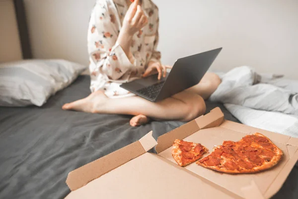 Woman in a pajama works on a laptop in bed and eats a pizza delivery.girl sits with a laptop on her feet at home in bed and eats fast food.Box of pizza pieces on the background of a girl with a laptop