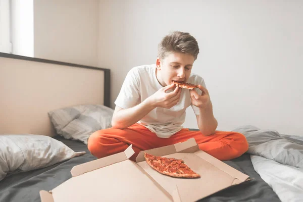 若い男は寝室でベッドの上に座っている 刺されボックスからピザの一個目を閉じて食べて 喜びです 朝食の配信からピザを食べています ファーストフード — ストック写真