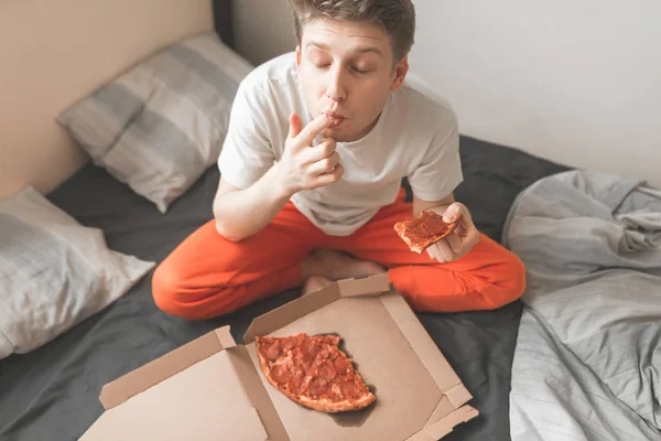 朝はピザのボックスに座っている ピザのピースの手を握り 味に彼の指をなめる若い男 男はベッドの午前中にピザを食べる 食品配送のコンセプト — ストック写真