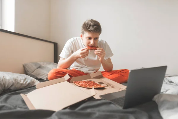 Young happy man is sitting on the bed with a box of pizza, uses a laptop and eats a piece of pizza. Boy sit on the bed, watching movies on a laptop and eating delivery pizza.