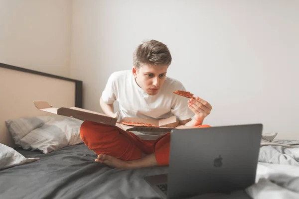 Young attractive man in a white t-shirt sitting at home, eating a piece of pizza and watching movies on a laptop. Teenager resting at home with a laptop and a delicious pizza delivery