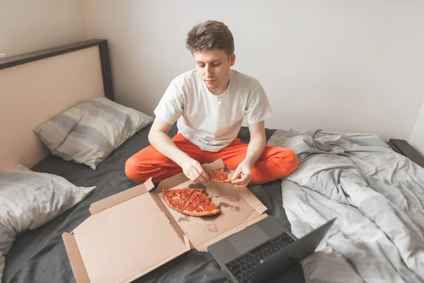 ベッドでファーストフードを食べて ラップトップを使用して少年の肖像画 ピザや ノート パソコンのボックスが付いているベッドに座ってる男 朝食とビデオを見て — ストック写真