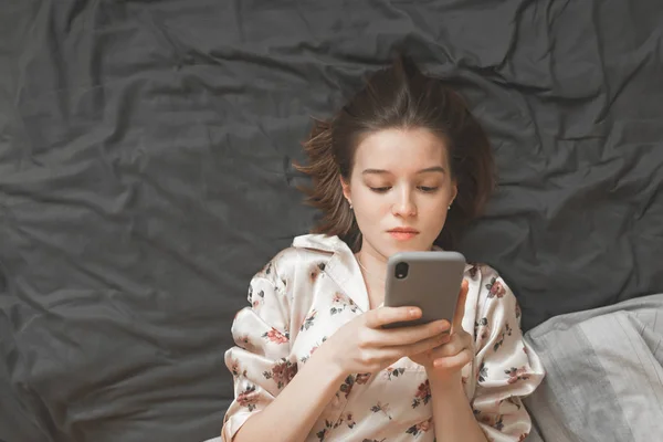 Portrait Charming Girl Pajamas Lying Bedside Table Looking Smartphone Screen — Stock Photo, Image