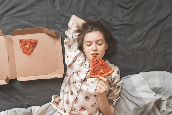 美しい少女の肖像パジャマに身を包んだ 目を閉じながらピザのピースを刺され 喜びを取得します 甘い女の子の自宅で安静時 ベッドでピザを食べて それを楽しんでします トップ ビュー — ストック写真