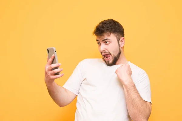 Joyful man wears a white T-shirt, looks at the smartphone and re — Stock Photo, Image