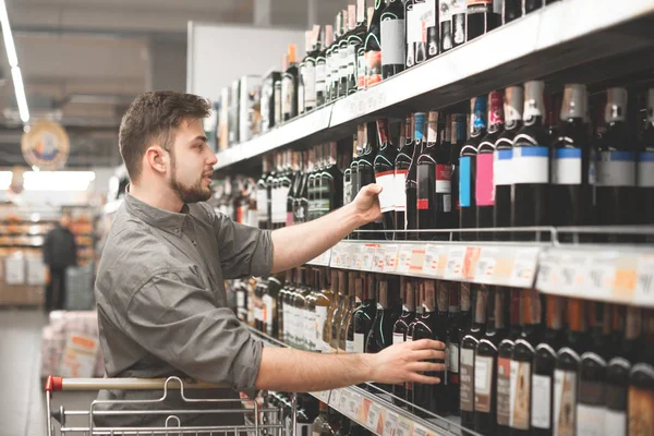 El hombre feliz con barba selecciona el vino en un supermercado, se para en los estantes con botellas y toma el vino con la mano. Hombre comprador toma el vino de los estantes del supermercado y sonríe . — Foto de Stock