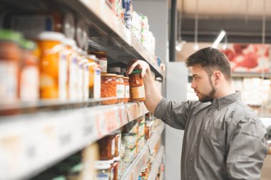 Adam bir süpermarket rafında konserve sebze seçer, bir kutu baklagiller tutar ve bir etiket okur. Alıcı bakkalda konserve sebze seçer. Boşaltmak