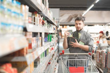 Mutlu adam bir süpermarket koridorda bir sepeti ile duruyor, bir indirimli olarak elinde birçok paket tutan, kamera görünüyor ve gülümsüyor.? bir alışveriş sepeti koymak raflardan çay birçok paket aldı