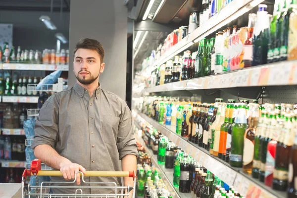 Retrato de un hombre con un carro, parado en el departamento de alcohol del supermercado cerca de los estantes con botellas de cerveza. Hombre comprador en el departamento de cerveza en el supermercado. Concepto de compras . — Foto de Stock