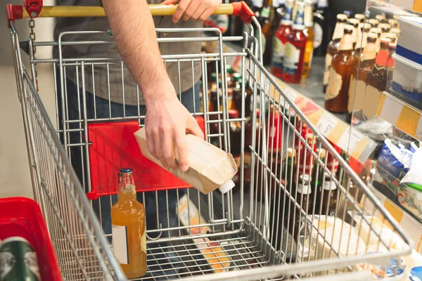Man's hand puts the products in an empty cart. Buyer makes purchases in a supermarket. Shopping in a supermarket concept. Buys drinks. Hands and cart close-up. Man buys alcohol — Stock Photo, Image