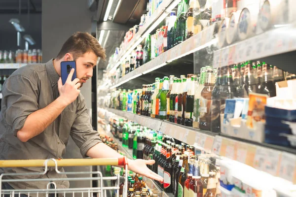 El hombre de negocios se para con un carrito en un supermercado, elige una botella de cerveza en los estantes en el departamento de alcohol y llama al teléfono. El hombre le pide a un amigo por teléfono que tome una cerveza en la tienda. — Foto de Stock